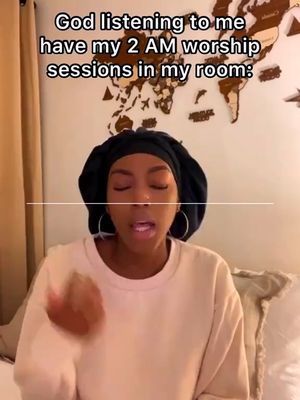 A woman wearing a black head covering is centered in a selfie-style shot. The background includes a decorative wooden world map and a neutral-toned wall. The woman is speaking with her eyes closed and her mouth slightly open, with one hand raised in a gesture.
