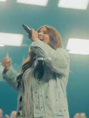 A woman with long, brown hair is singing into a microphone on a stage. The background is softly lit with cool tones, with a text overlay that says "GREAT IS".