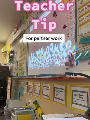 The image shows a classroom scene. Above the whiteboard, the text reads 'Teacher Tip' in pink bubble letters, with 'For partner work' below in a white box. Classroom walls are covered with posters and learning tools. A teacher's desk and a projector are visible.