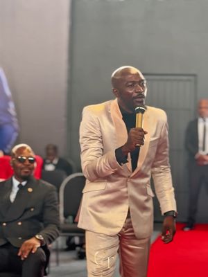 A man in a shimmering, gold suit speaks into a microphone, with a group of people in the background at a ceremony. The lighting is warm, with a red platform adding a focal point.