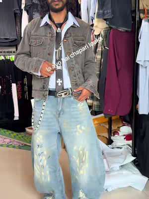 A man stands in a clothing store, dressed in a layered outfit. He wears a gray denim jacket over a collared white dress shirt and a black tie with cross and skull motifs. Light blue, wide-legged jeans and a silver belt buckle complete the look. The background displays clothing racks.