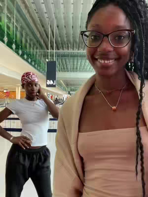 A selfie-style video cover image featuring two young women in an airport or public space. The foreground woman, with glasses and long dark hair, smiles directly at the camera. Behind her, another woman stands and poses, also looking toward the camera. The background shows a modern building interior with structural elements and neutral colors.