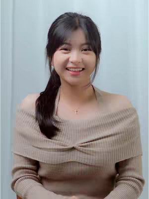 A young woman with black hair pulled back in a ponytail smiles at the camera. She wears a light brown, off-the-shoulder sweater. The background is a plain, light-colored wall.