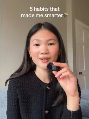 A young woman with long black hair is positioned in front of a blurred background. She is wearing a black, button-down sweater and holds a small microphone near her mouth, appearing to be speaking. Above her, a text overlay reads '5 habits that made me smarter' in a white font. The composition is a medium shot, focusing on her face as she speaks.