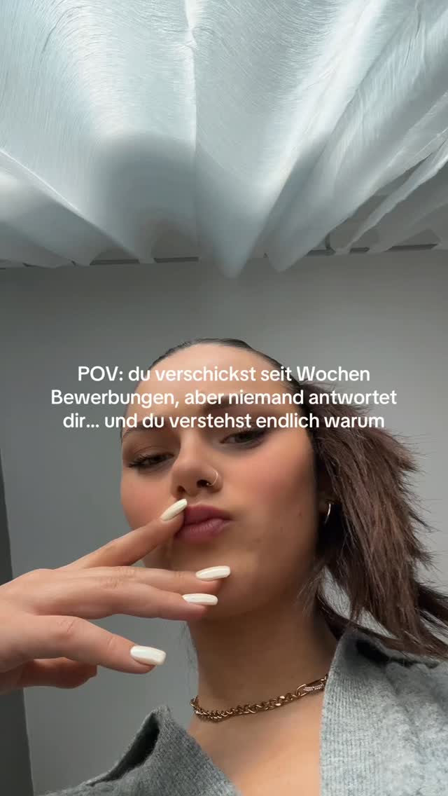 A selfie-style shot featuring a young woman looking at the camera with a slightly puckered expression. She has light skin, a nose ring, and short dark hair. Her left hand is raised, with the index finger touching her lips. The background includes white, textured surfaces, possibly fabric or a wall.