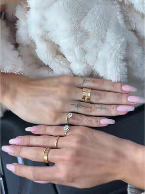 Two hands with long, pink nail polish are displayed, each wearing multiple gold rings. The hands rest against a shaggy, white, fluffy background. The top hand makes stars tattoo on finger.