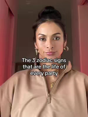 A woman with dark hair pulled back in a bun stands in a hallway, facing the camera. She is centered in the frame and wears a tan-colored zip-up, with simple gold earrings and necklace. The background is a blurred hallway with a pinkish-red hue, and additional text is on the screen.