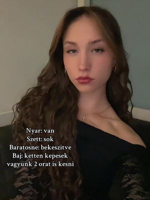 A young woman is centered in the frame, with long, wavy brown hair. She is wearing a black top and a gold necklace. Text overlays are present, written in Hungarian, on the left side of the image against a neutral background. The lighting appears soft, and the overall composition is a selfie-style shot.