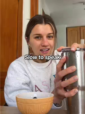 A woman with light brown hair and a white sweatshirt is looking at the camera with a slight smile. She is holding a stainless steel tumbler in her right hand and a bowl in her left hand. The scene appears to be indoors, with a wooden cabinet in the background.