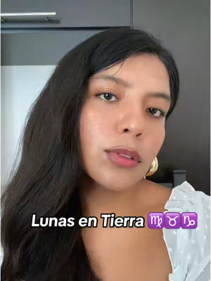 A woman with long black hair and large gold hoop earrings looks directly at the camera. She is wearing a white top, and the background is out of focus. Text overlaid on the image reads 'Lunas en Tierra' with zodiac symbols for Taurus, Virgo, and Capricorn.
