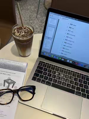 A photograph taken from an overhead perspective showcases a Macbook laptop open to a study page, a cup of iced coffee with a straw, a pair of glasses, and a notebook on a light-colored surface. The coffee cup is on the upper left, with the macbook and study materials angled towards the right. The overall aesthetic is casual and modern, typical of a study setting. The items suggest a setting intended for study or work.