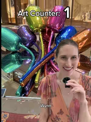A woman smiles at the camera in front of a colorful sculpture resembling balloon-like tulips. Above her head, text reads 'Art Counter: 1'. The setting appears to be a modern, well-lit space, likely a public area like a hotel lobby.