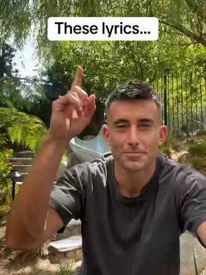 A man with short, dark hair and a short beard, wearing a dark t-shirt, gestures upwards with his right index finger. He is in a well-lit outdoor setting with lush green foliage in the background. A text overlay at the top reads "These lyrics..."