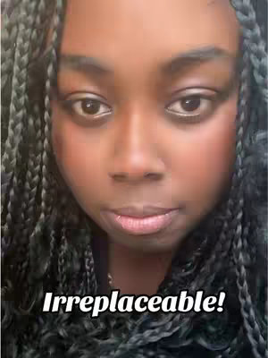 A close-up shot of a person's face with dark skin, framed from the chest up. Braided black hair surrounds the face. The person has dark eyes looking towards the viewer, and a neutral expression. They have lightly applied blush and lipstick. The background is out of focus, but appears to be a light-toned wall.