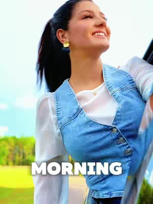A woman with dark hair pulled back in a ponytail, wearing a denim vest over a white top, looks upwards with a joyful expression. The background shows a green, blurred landscape, suggesting an outdoor setting with sunlight and a warm color.