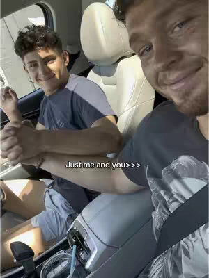 A selfie taken from inside a car featuring two men, one taking the photo and the other smiling at the camera. They are holding hands, conveying a sense of relationship. The setting is a car interior, and the color palette is natural, with the focus on the individuals.