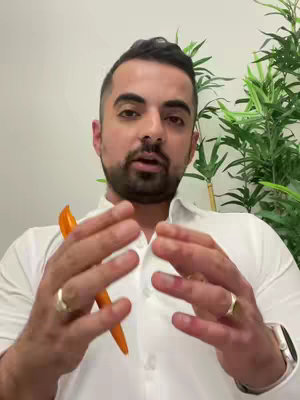 A man with a beard is speaking to the camera, with both hands open in a gesture. He is wearing a white shirt and two rings, one in each hand. Behind him is a light wall and a leafy green plant.
