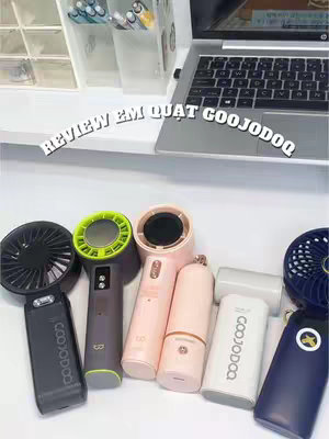 The image showcases a collection of handheld fans arranged on a white surface, with a laptop in the background. The fans come in various colors and designs. Above the fans, a text overlay reads 'REVIEW EM QUAT GOOJODOQ'. The overall composition is clean and product-focused.