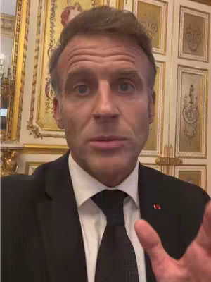 A medium shot of Emmanuel Macron, the President of France, looking directly at the camera and speaking. He's wearing a black suit, white shirt, and a dark tie along with a lapel pin. The background is a room with gold trim and details, suggesting a formal setting. His expression is serious, and he seems to be making a statement or addressing a question.