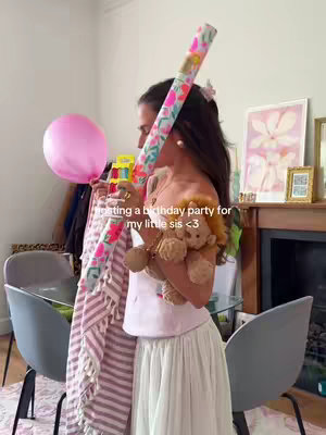 A young woman stands in a well-lit room, holding a pink balloon and a gift, possibly wrapping paper. She appears to be setting up for a birthday party. The background includes a fireplace and artwork, suggesting a home environment.