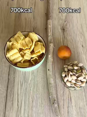 The image presents a wooden surface on which three items are arranged. On the left, a bowl filled with yellow, crispy chips is positioned next to an object that resembles a beige twig. On the right, a small glass bowl contains pistachios and a single orange fruit, likely a mandarin or a small nectarine. Text indicating '700kcal' is placed above the chips and the pistachios.