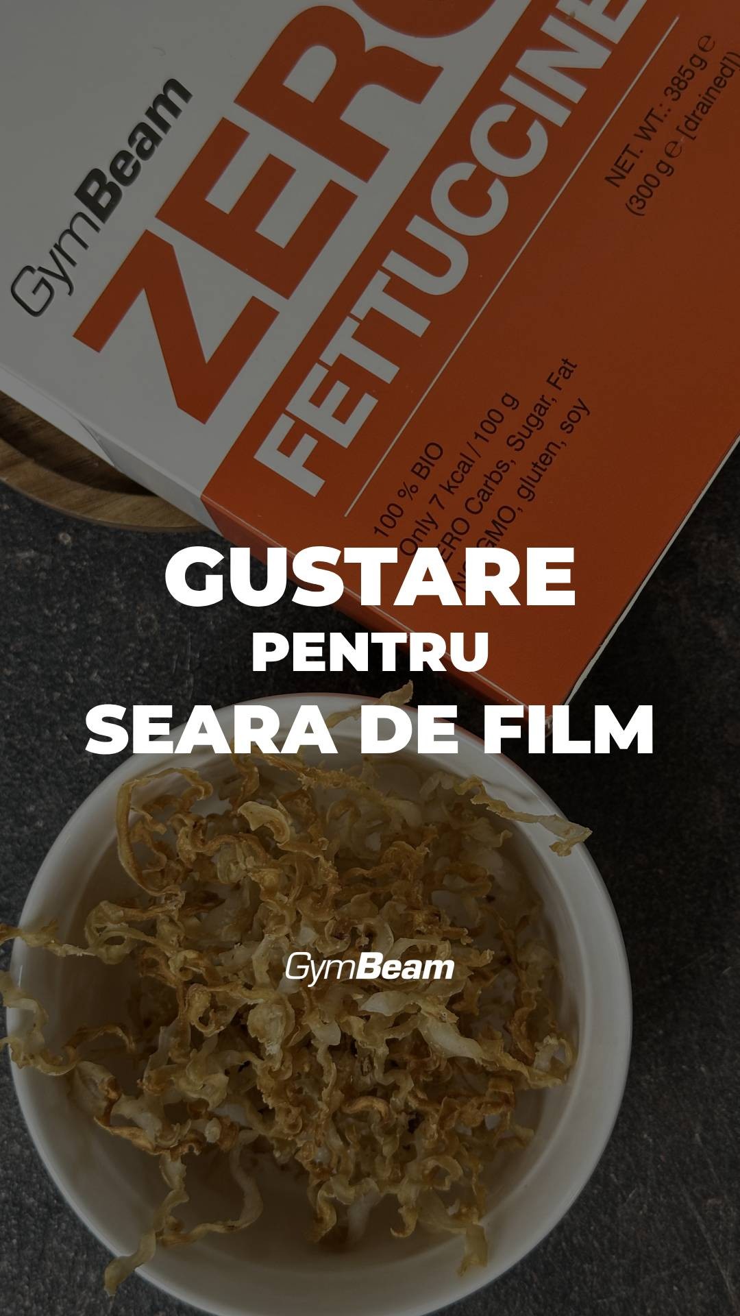 The image features a top-down view of a bowl filled with what appears to be crispy, golden-brown fettuccine. Below the bowl is a dark, textured surface. The background presents an open box with the GymBeam brand displayed prominently with orange and white colors.