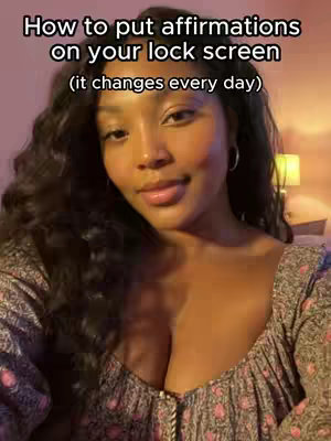 A woman with long, wavy black hair is centered in the frame. She is wearing a top with a floral print. The background is subtly blurred, featuring warm lighting, suggesting an indoor setting. Above her, text in white font reads 'How to put affirmations on your lock screen,' followed by '(it changes every day)' in a smaller font also in white.