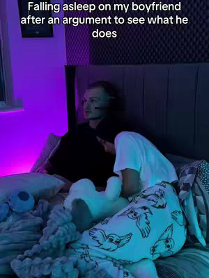 The image shows a couple in bed. One person is lying face down, tucked under a blanket, while the other is sitting up and looking at the person lying down. The room's ambience is enhanced by purple lighting emitting from the left side of the image.