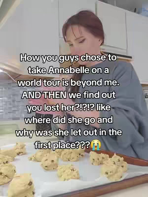 The cover image shows a woman in a kitchen setting. She is holding a baking tray with several chocolate chip cookie dough balls on it. Text is overlaid on the image, conveying a humorous reaction to a situation involving the Annabelle doll. The background shows a kitchen with appliances (oven, cabinet). The scene is well lit, making the colors appear natural.