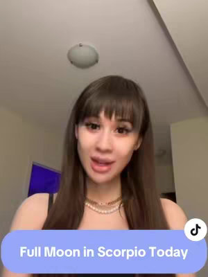 A woman with long brown hair and bangs is centered in the frame. She is looking towards the camera with a subtle smile. The background is blurred, and a text overlay in a blue bubble at the bottom reads, "Full Moon in Scorpio Today."