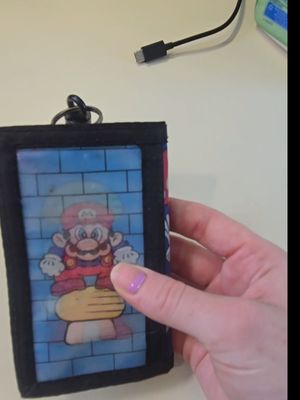 A hand, with polished nails, clasps a black and blue wallet that has an image of the character Mario from the video game Super Mario, as if the image were printed on a see-through piece of plastic on the wallet. A USB cable is visible in the top right.