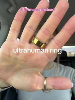 A close-up shot focuses on a hand displaying rings. The hand, featuring a gold ring on the middle finger and another on the index finger, is set against a blurred background, possibly inside a car. Text overlays read "off Amazon instead" above and "ultrahuman ring" below the hand, highlighting the products.