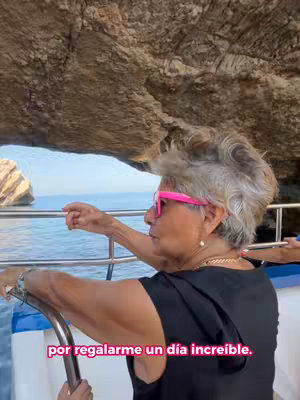 A medium shot shows an older woman on a boat with her arm resting on the railing. She wears pink framed sunglasses, a black top, and a gold chain necklace. The image has the texture of rock formations in the background, and the water and sky are also visible.