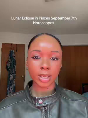A close-up shot featuring a Black woman with smooth skin, highlighted with a red filter. She is looking directly at the camera with a neutral expression, her dark hair pulled back. She is wearing a leather jacket, and her eye makeup is well-defined with sharp eyeliner and some eyeshadow. The background is slightly blurred, but there are hints of a room.