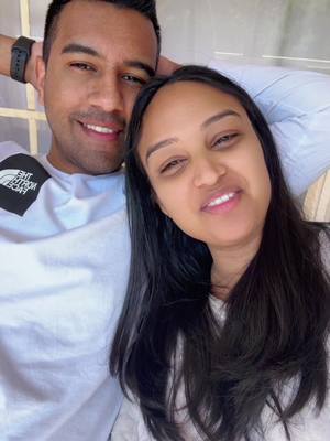 A selfie of a couple, close up, smiling at the camera. The man is on the left, wearing a white t-shirt and a black watch. The woman is on the right with dark hair and smiling. The background is blurred.
