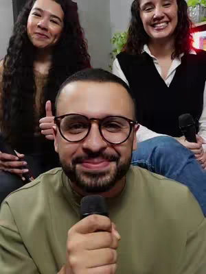 A selfie-style video cover featuring three people. In the foreground, a man with glasses holds a microphone and smiles at the camera. Behind him and to the left, a woman with dark curly hair smiles, raising her thumb. To the right, another woman with curly hair also smiles, accompanied by a microphone held to her lap. The background suggests an indoor setting, possibly resembling a studio.