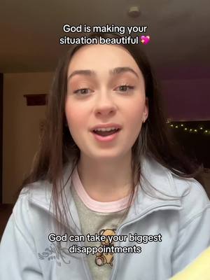 A young woman with long brown hair is centered in the frame, smiling and speaking. Text overlays read "God is making your situation beautiful" at the top and "God can take your biggest disappointments" at the bottom. She's wearing a light blue jacket over a grey shirt with a teddy bear graphic, and the background fades into neutral tones.