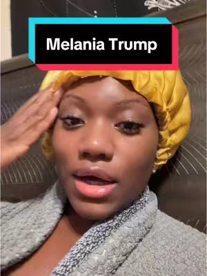 A selfie-style shot featuring a young woman with dark skin, looking directly at the camera with a slight smile. She's wearing a yellow head covering. Overlay text says, "Melania Trump." The background includes a dark, patterned surface, likely a bed or a wall.