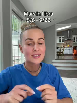 A woman with blonde hair in a topknot is centered, facing the camera and speaking. Behind her is her home interior, with a doorway leading to a kitchen. Text at the top reads 'Mars in Libra 8/6 - 9/22'.