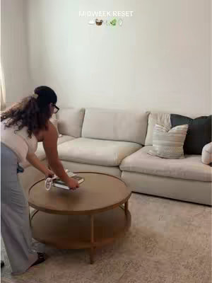A woman stands in a living room, arranging items on a coffee table. The room has a beige sectional sofa, round wooden coffee table, and neutral-toned rug. The woman is wearing glasses, a light-colored top, and grey pants. The text overlay 'MIDWEEK RESET' is at the top of the image with some emojis.