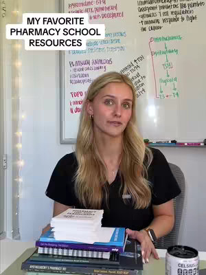 A woman is seated, holding a collection of books with a white board behind her. The board is filled with handwritten notes and diagrams related to pharmacology. The woman is facing the camera and looking towards the viewers. A stack of books and a can of Celsius drink are on the desk, signaling a study or work context.