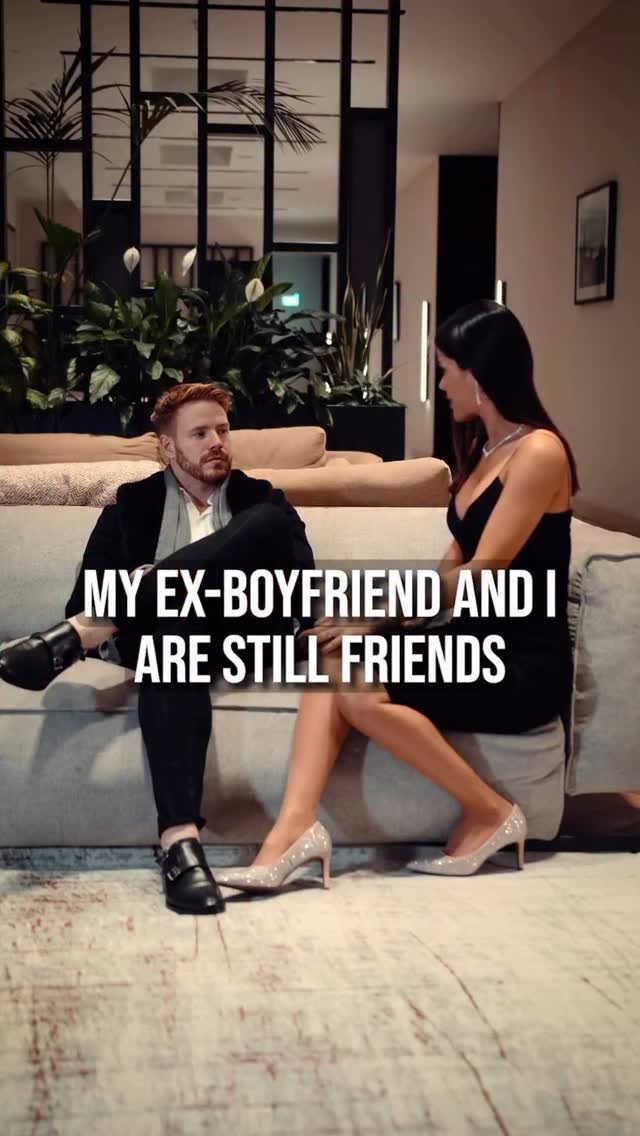 A man and a woman are seated on a couch in a modern, well-lit space. The man, with red hair and a short beard, is leaning back, wearing a black jacket and dark pants. The woman, with dark hair, is sitting next to him in a little black dress with a sparkling shoes. Text overlayed on the image reads 'MY EX-BOYFRIEND AND I ARE STILL FRIENDS'. The background includes a window, plants, and a decorative panel.