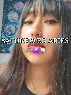 A selfie-style shot with a woman as the central figure. She has long, dark hair with bangs, dark eyeliner, and a nose ring, and her lips are lightly colored. The background is a bit soft but features a patterned wall. Overlaid on her face is the text 'SATURNO EN ARIES' in white against a black background. Below this text, there are various emojis.