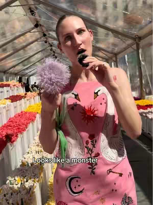 A woman in a pink dress with a swan design stands in front of a flower market, holding a purple pom-pom-like object and an object that she appears to be eating or about to eat. The background shows rows of colorful flowers and a transparent structure overhead.