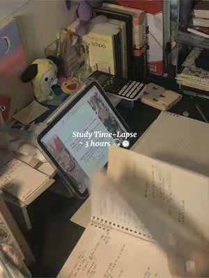 A cluttered desk with a laptop, books, and stationery. The laptop screen displays content while pages of a notebook are being turned in the foreground.