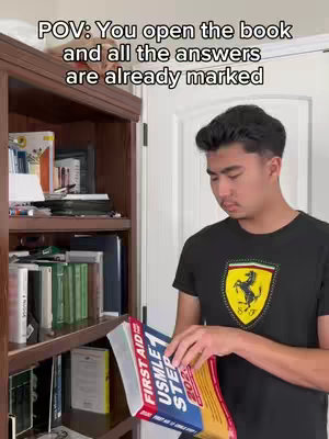 A young man stands in front of a bookshelf, holding a textbook titled 'First Aid for the USMLE Step 1 2023'. He's wearing a black t-shirt featuring a Ferrari logo. Overlay text in a bubble reads 'POV: You open the book and all the answers are already marked'.