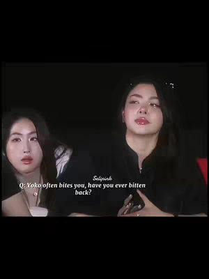 Two Asian women are seated in front of a dark background, centered in the frame. The woman on the right is the primary focus, with long dark hair and a black top, her gaze slightly upward. The second woman, positioned to the left, is also Asian with dark hair and a neutral expression. A text overlay in the image reads, "Q: Yoko often bites you, have you ever bitten back?" adding an element of curiosity. The overall composition is simple, focused on the two subjects and the interaction.