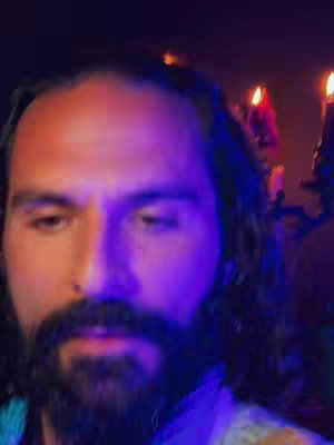 A close-up shot of a man's face with a thick mustache and beard, looking slightly downward. The lighting is a mix of warm light on his face and cool purple light in the background, creating a dramatic effect. Behind him, blurred candles add to the ambiance.
