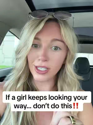 A woman with blonde hair is inside a car. She is wearing sunglasses atop her head and is looking at the camera and speaking. There is a white text box with black text overlayed at the bottom of the screen.