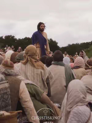A wide shot shows a man in ancient attire addressing a crowd. He stands on a slight rise amid a sea of people. The man, presumably Jesus, wears a tan robe and a dark blue shawl. The crowd is dressed in similar period clothing, with many wearing head coverings. The background is a blurred mix of greenery and a cloudy sky. The overall effect is one of historical depiction and religious significance.